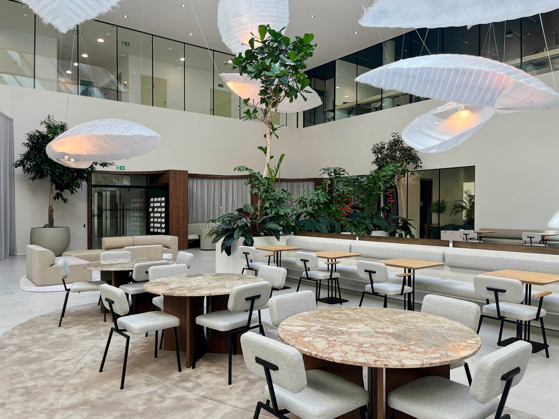 Grand lounge d'affaires lumineux avec tables rondes en marbre, chaises claires, plantes et suspensions en tissu blanc