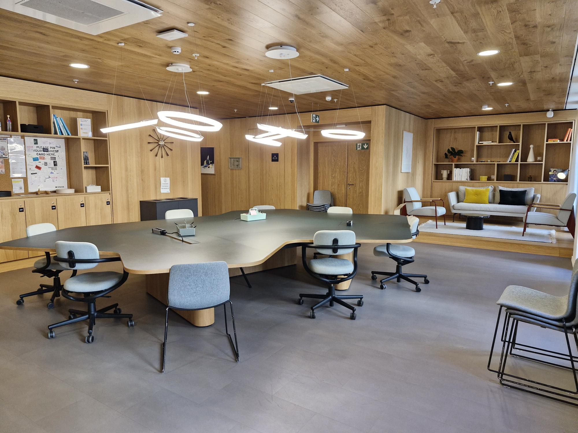 Espace de travail partagé avec grande table, chaises grises, plafond en bois, suspensions lumineuses et coin salon